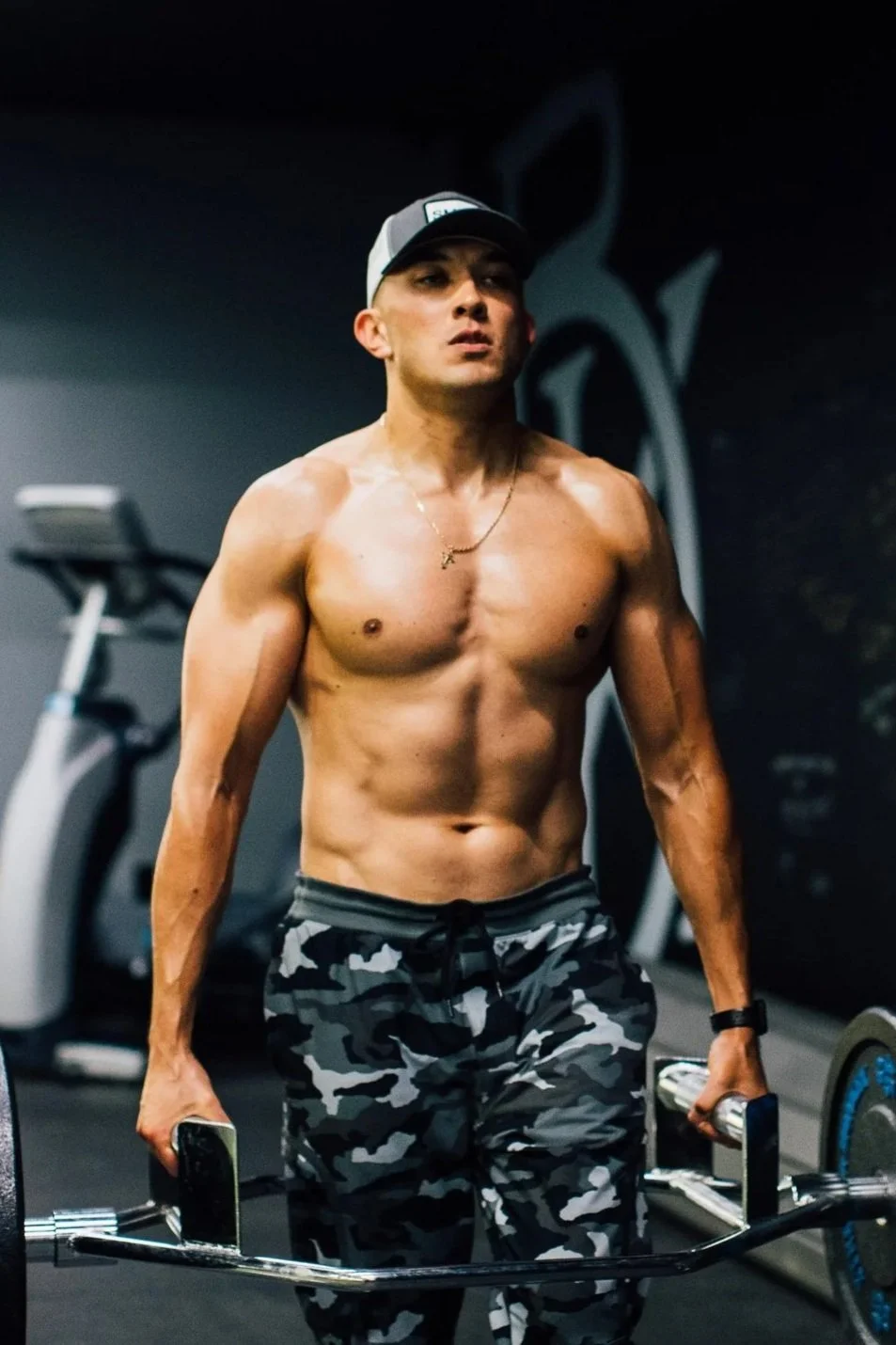 A shirtless young man with a cap, gold chain, and camo shorts lifting weights with dumbbells in a gym.