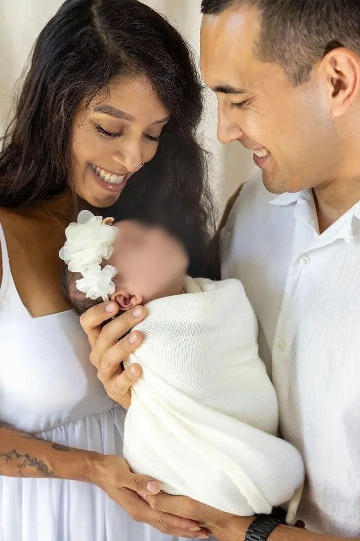 A happy family consisting of a smiling woman, a smiling man, and a baby wrapped in a white blanket and wearing a white flower headband, during a special moment in a home or studio setting.