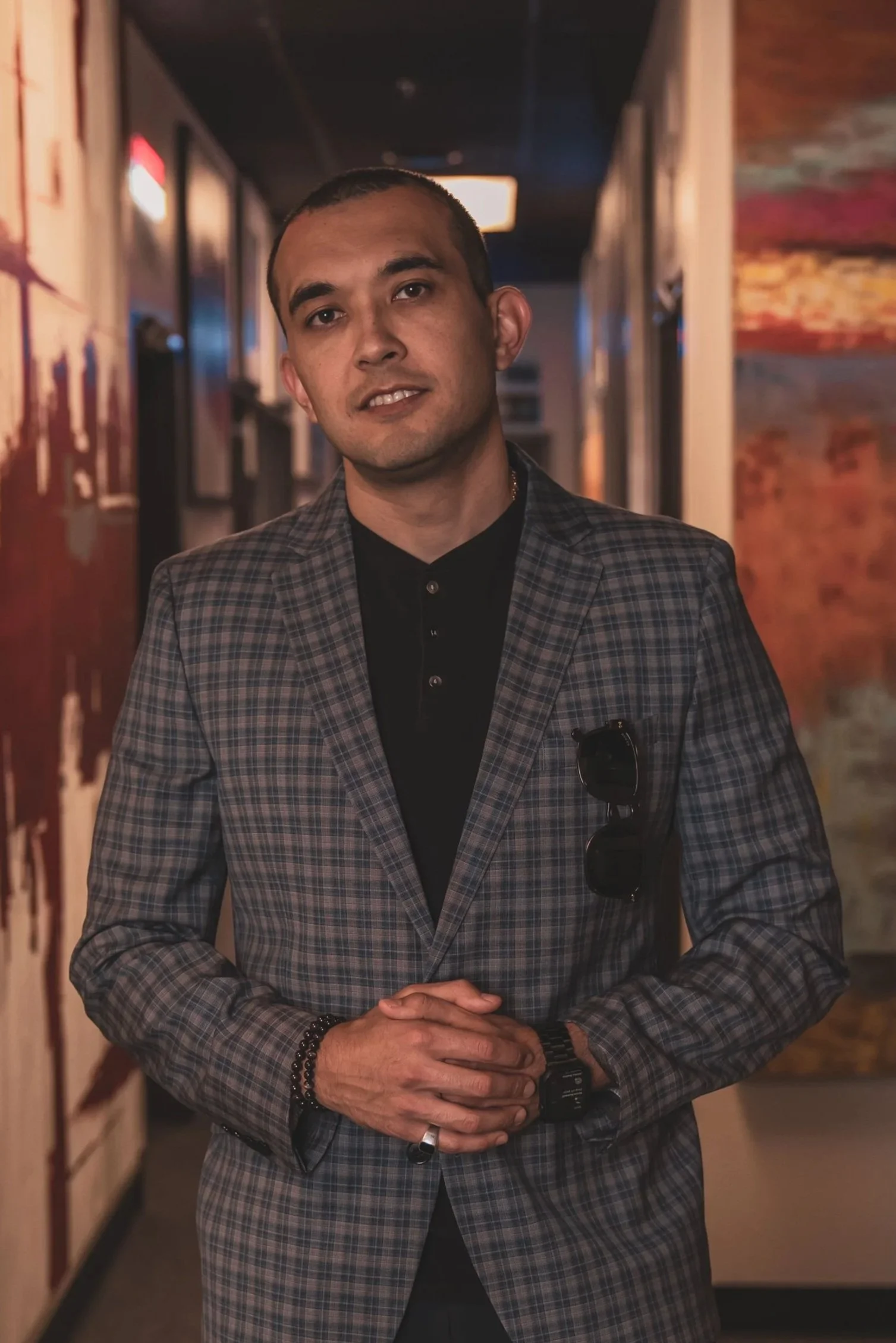 A man in a plaid blazer standing in a dimly lit hallway with artwork on the walls and his hands clasped in front of him.