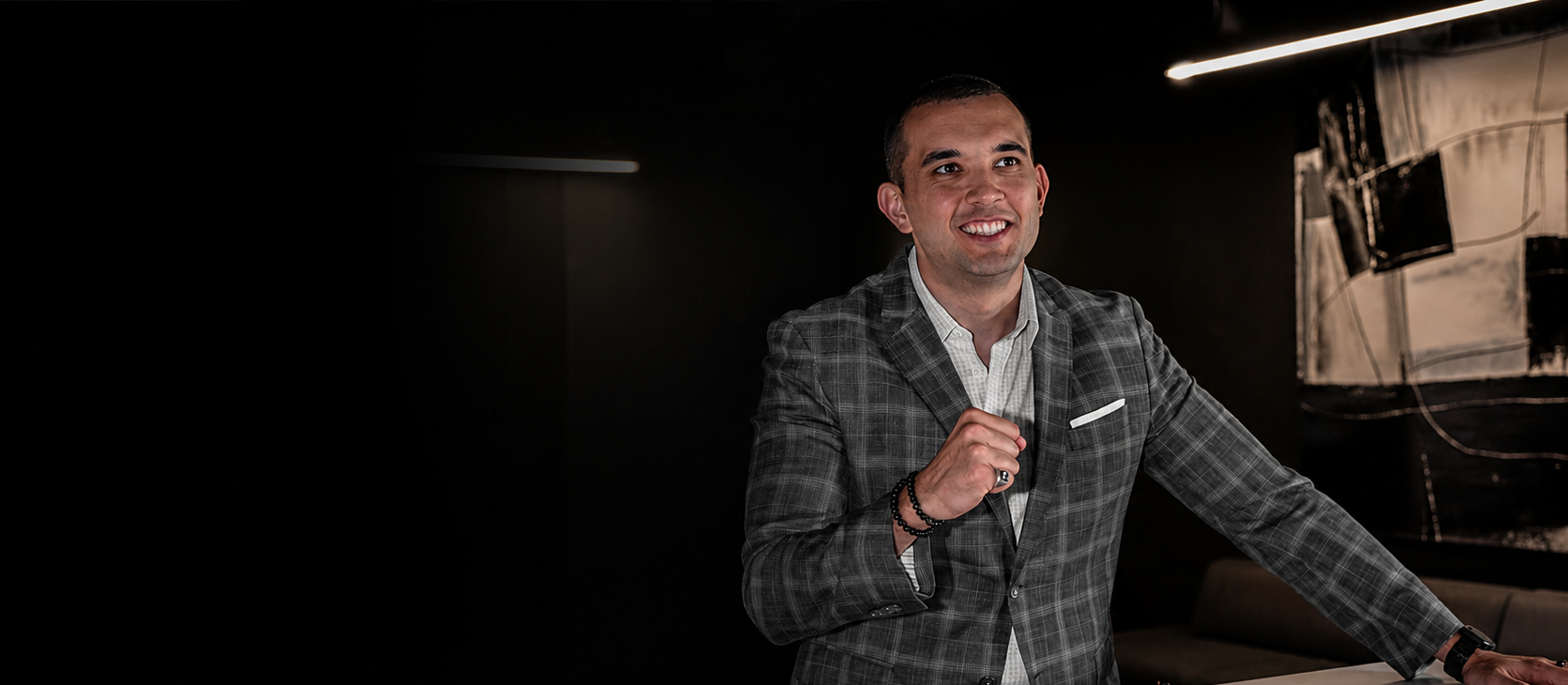 A smiling man in a checkered suit standing at a table in a dark room with modern lighting and abstract art on the wall.