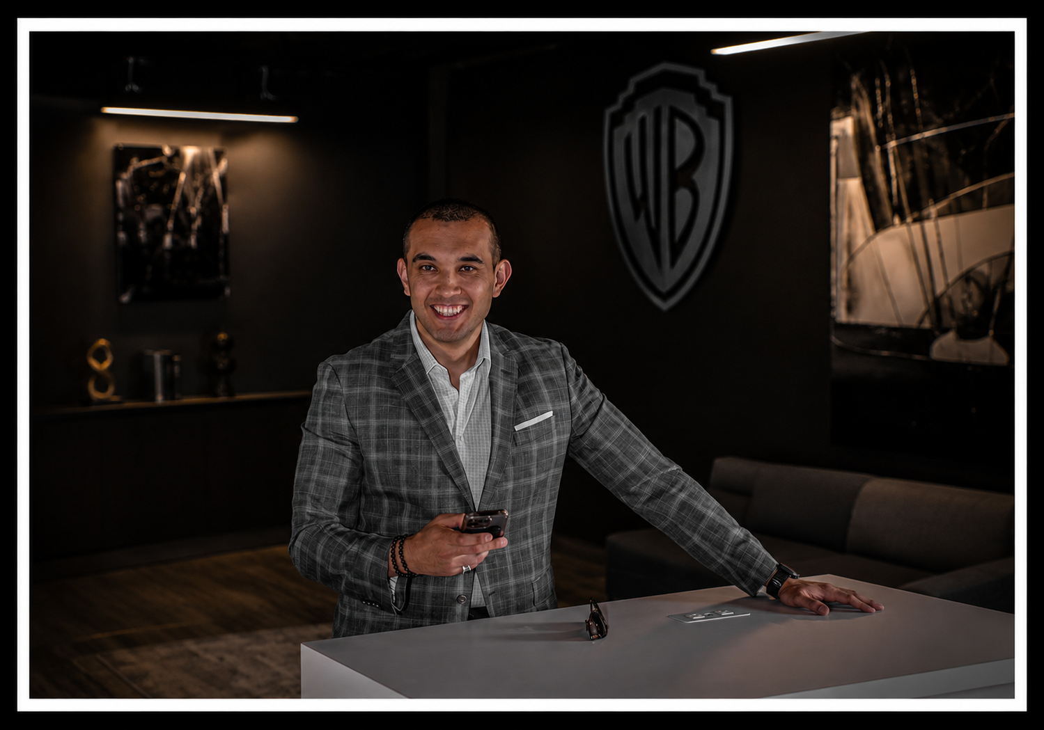 A smiling man in a gray checkered suit and white shirt standing at a modern reception desk in a dark, stylish office or lounge. He's holding a smartphone and has sunglasses on the desk in front of him. The background features a black wall with the Warner Bros. logo and abstract art.