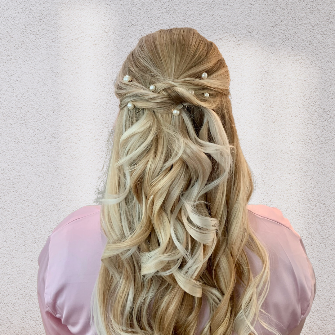 Back view of a woman with long, wavy blonde hair styled with small pearl hair accessories, wearing a light pink satin blouse against a white wall.