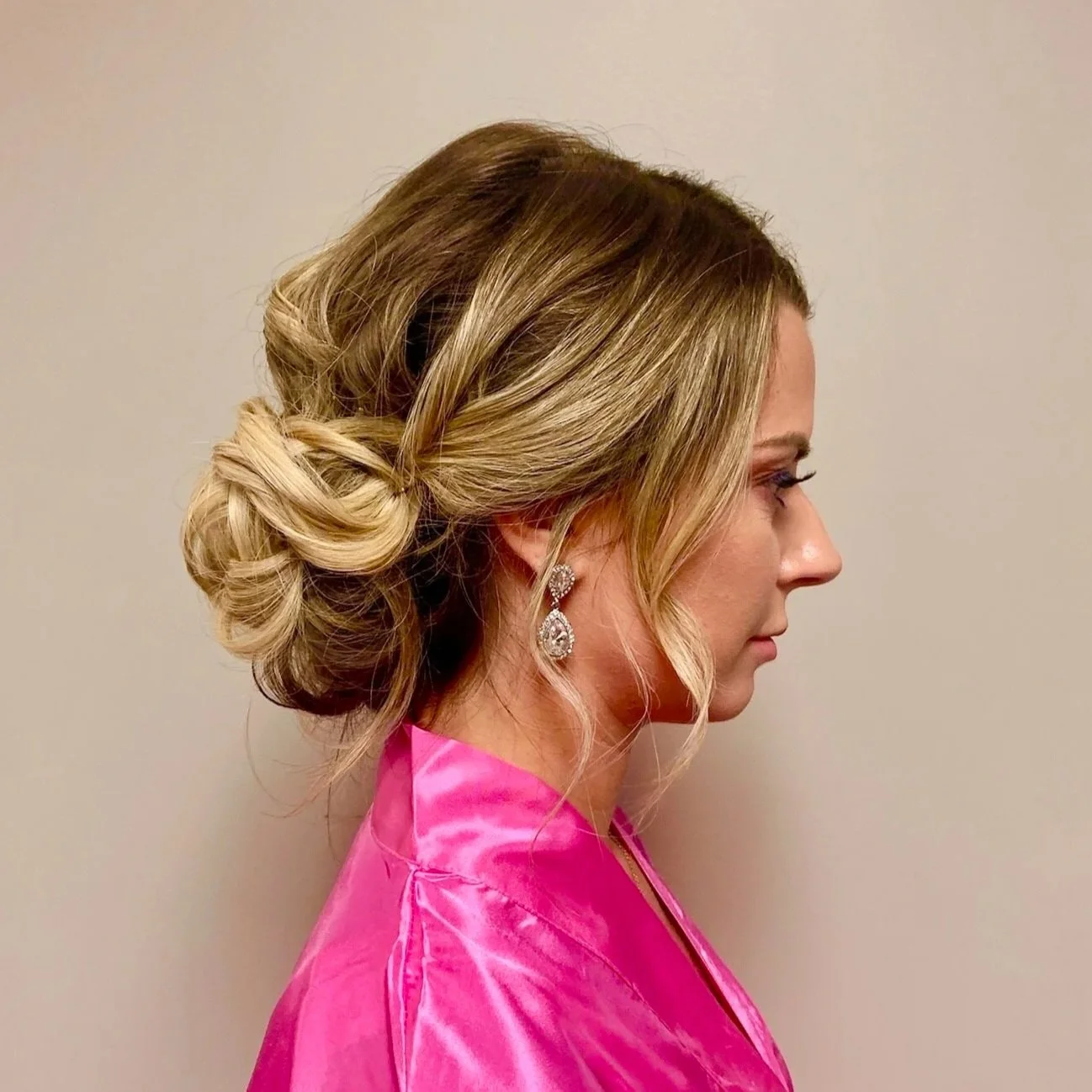 A woman with styled blonde hair in an elegant updo, wearing diamond earrings and a bright pink satin robe, facing sideways against a plain light gray background.