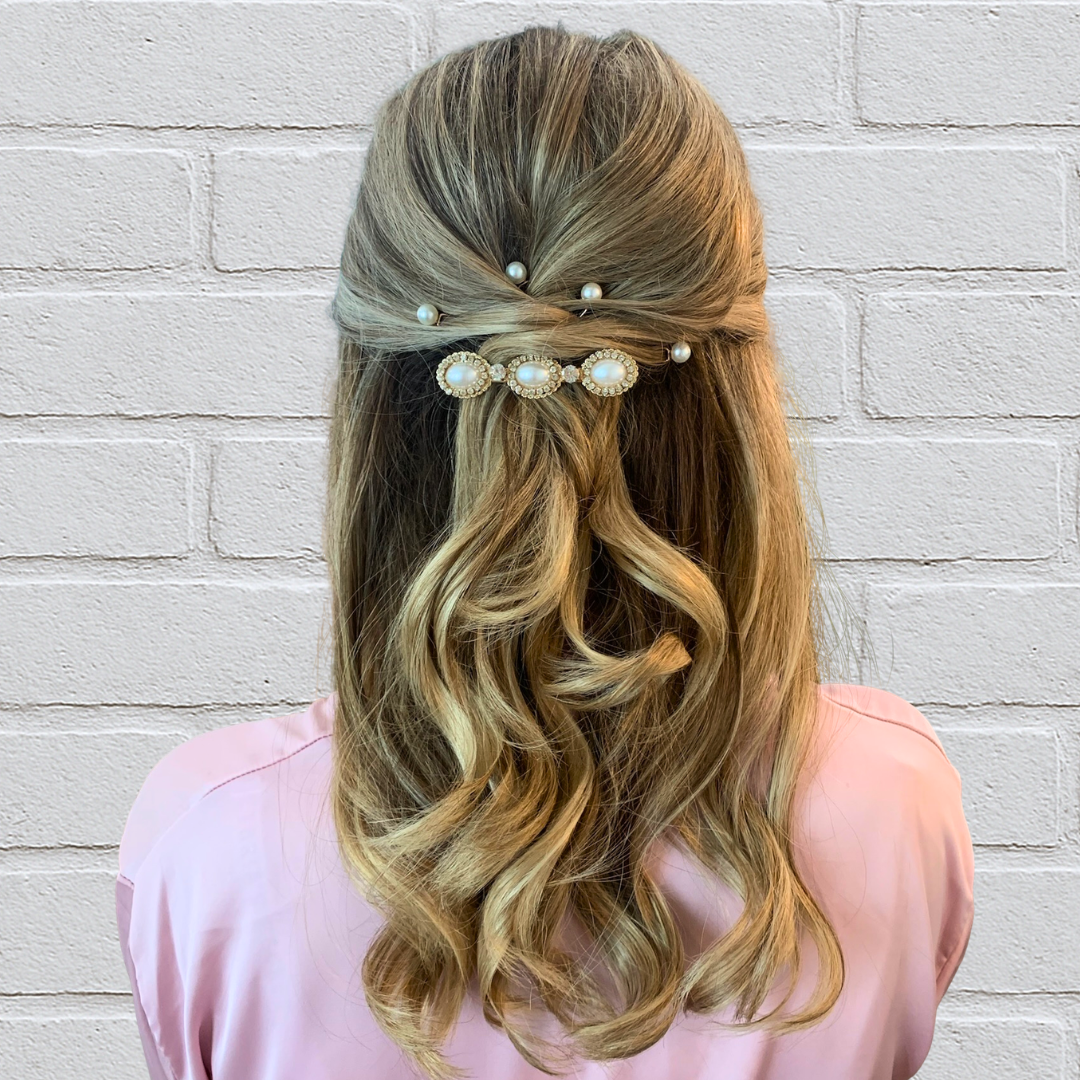 Back view of a woman with wavy blonde hair styled with pearl hairpins and decorative pearl hair clips, wearing a pink top, standing against a white brick wall.