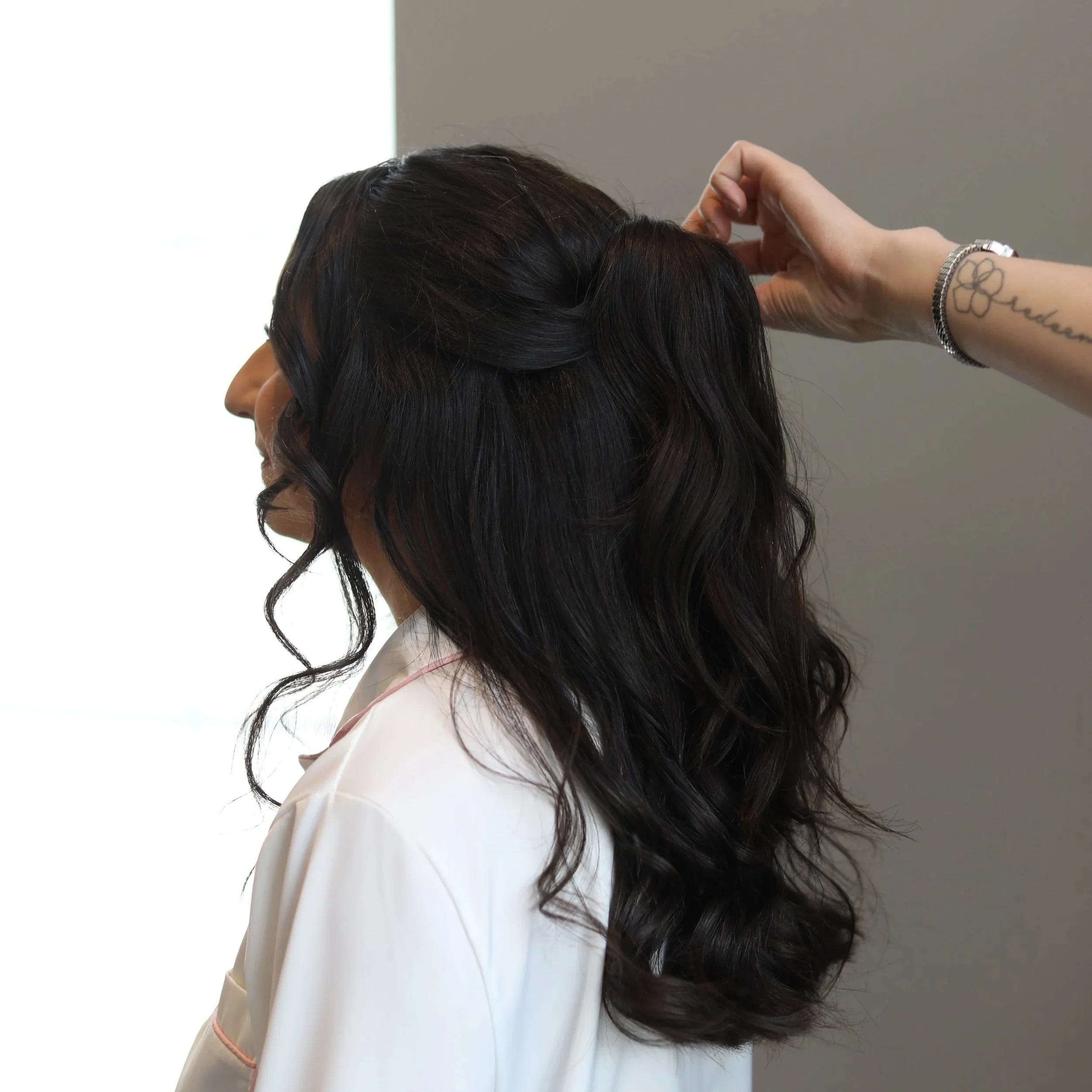 Close-up of a woman with long dark wavy hair, partially pulled back, wearing a white top, with a small tattoo of a flower and the word 'peace' on her left wrist.