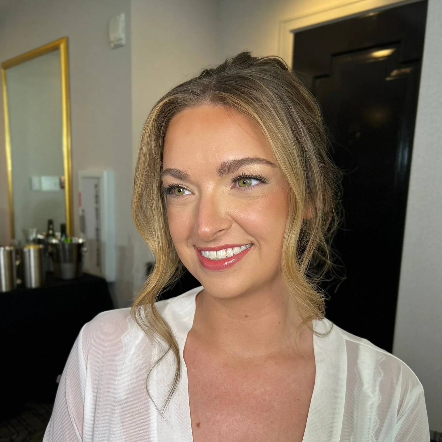 A woman with blonde hair styled in loose waves, smiling, wearing a white top, in an indoor setting likely at a salon or event.