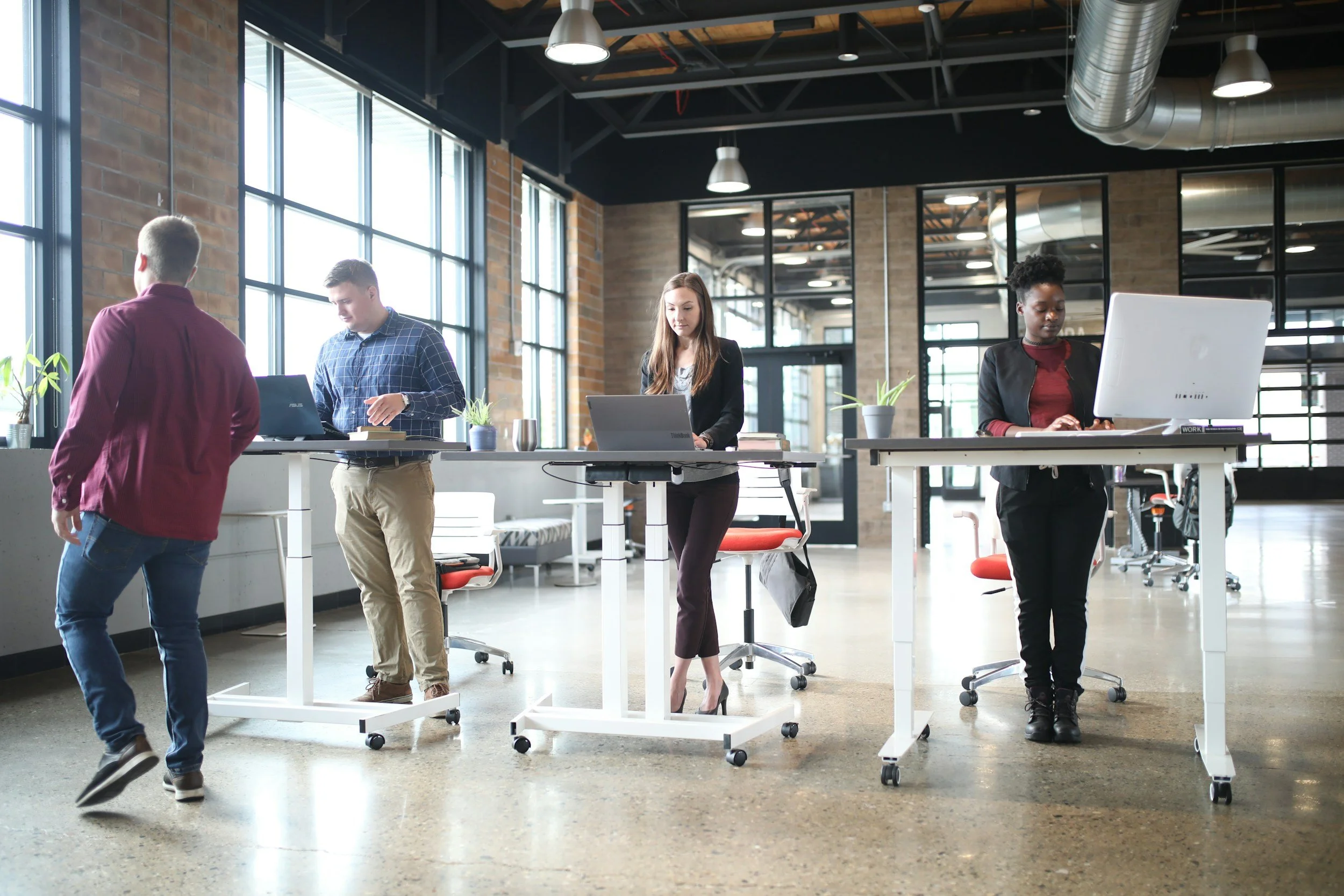 Personas trabajando en una oficina moderna con grandes ventanas, escritorios con computadoras portátiles y plantas decorativas, en un ambiente de espacio abierto con techo industrial y aire acondicionado.