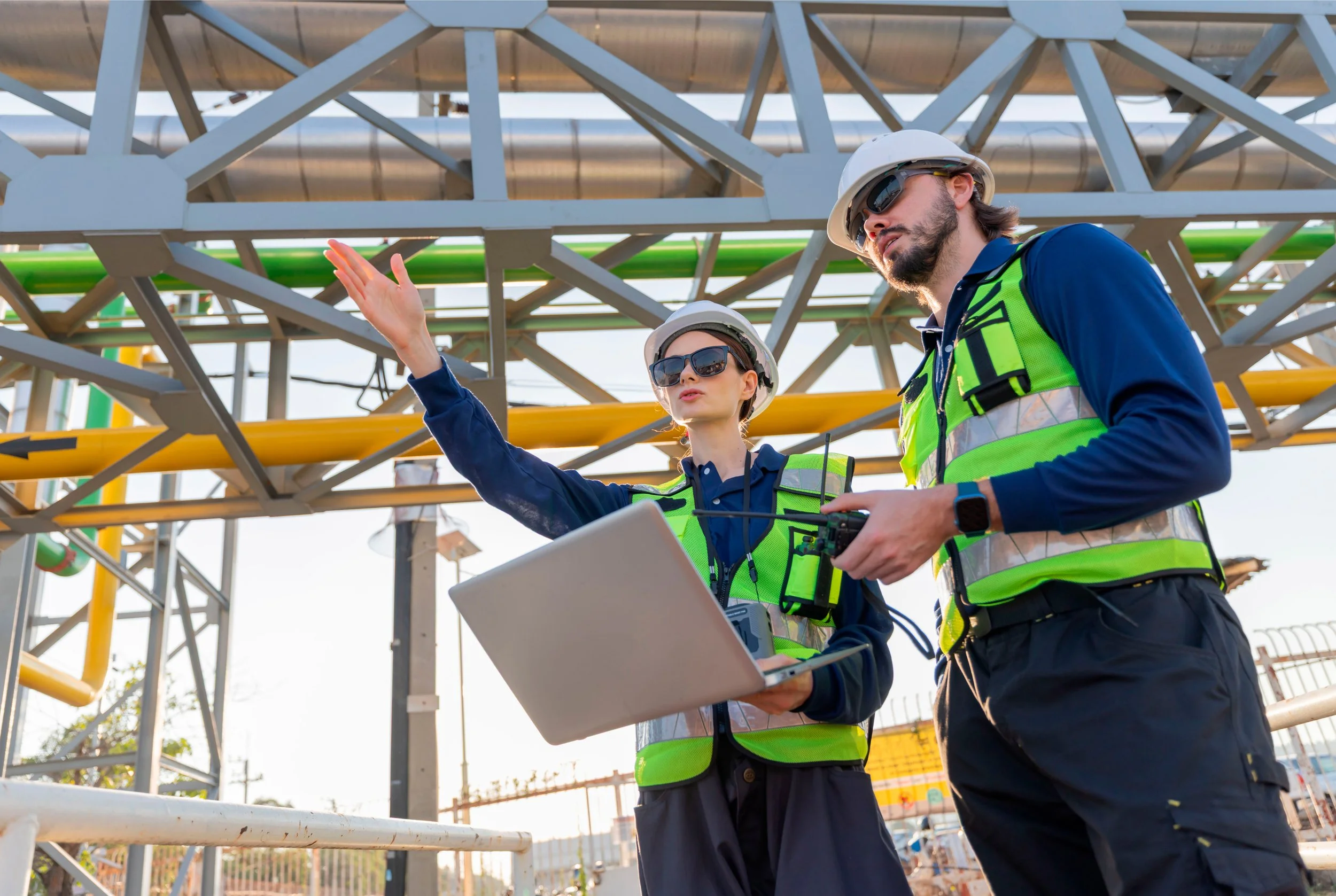 Dos técnicos en seguridad industrial usan cascos, gafas de sol y chalecos reflectantes mientras inspeccionan una estructura metálica y controlan un equipo portátil, en un entorno de construcción industrial.