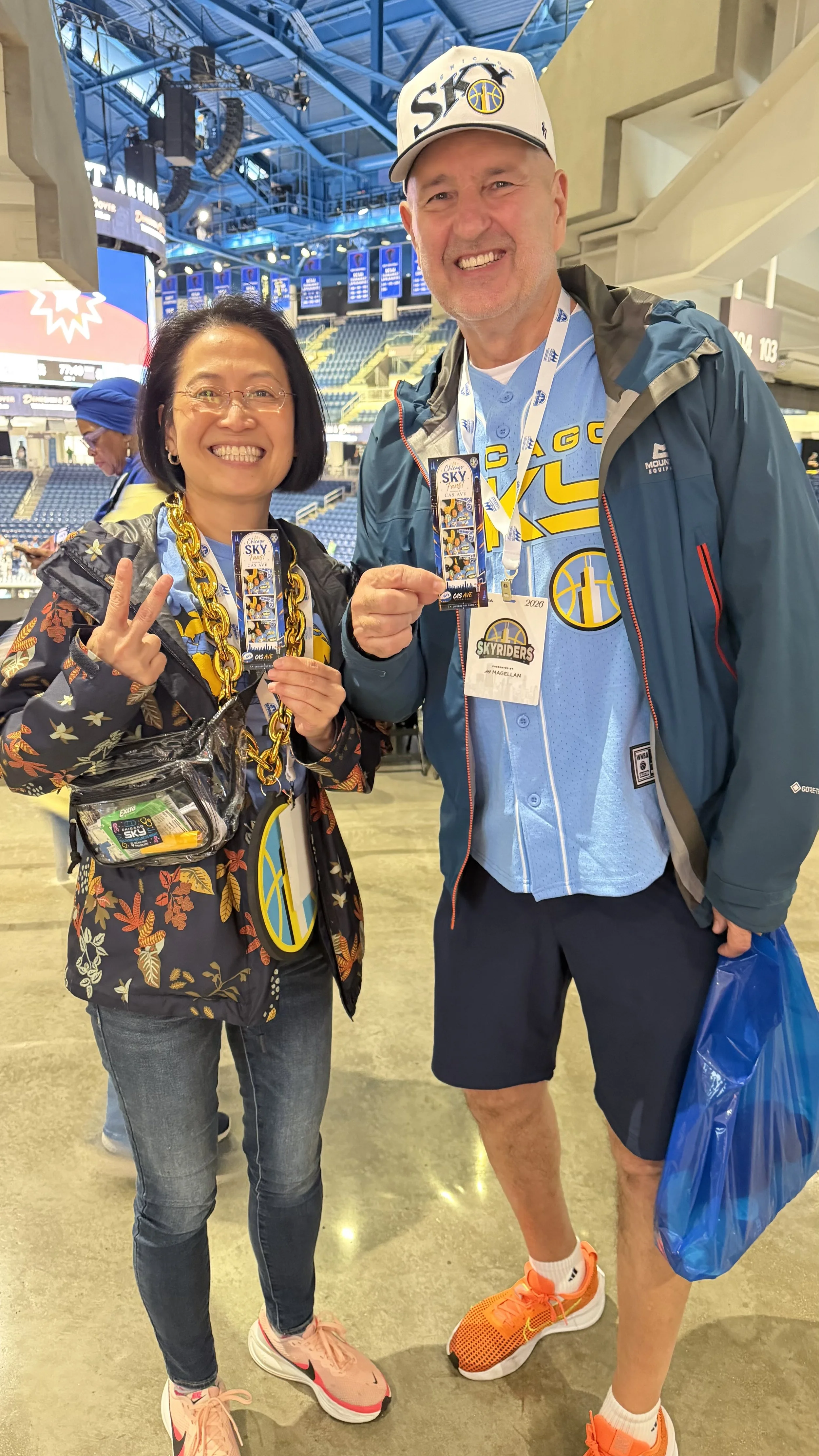 Smiling woman and man at a sports arena, wearing Chicago Sky team gear, holding tickets, with the arena's interior visible in the background.