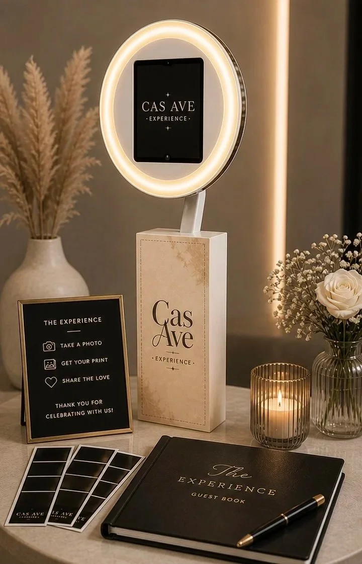 Decorative table display at Gas Ave Experience featuring a mirror with a light ring, a sign with instructions for guests, a guest book titled "The Experience," a pen, photo strips, a candle, a vase with white roses and baby's breath, and pampas grass in a large vase.