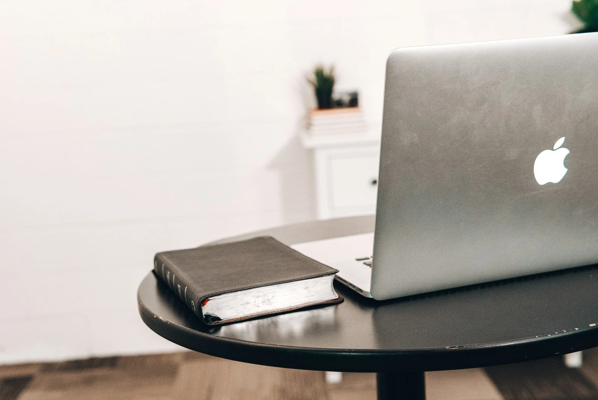 A closed black notebook with a leather cover resting on a dark round table next to a silver MacBook laptop in a bright room.