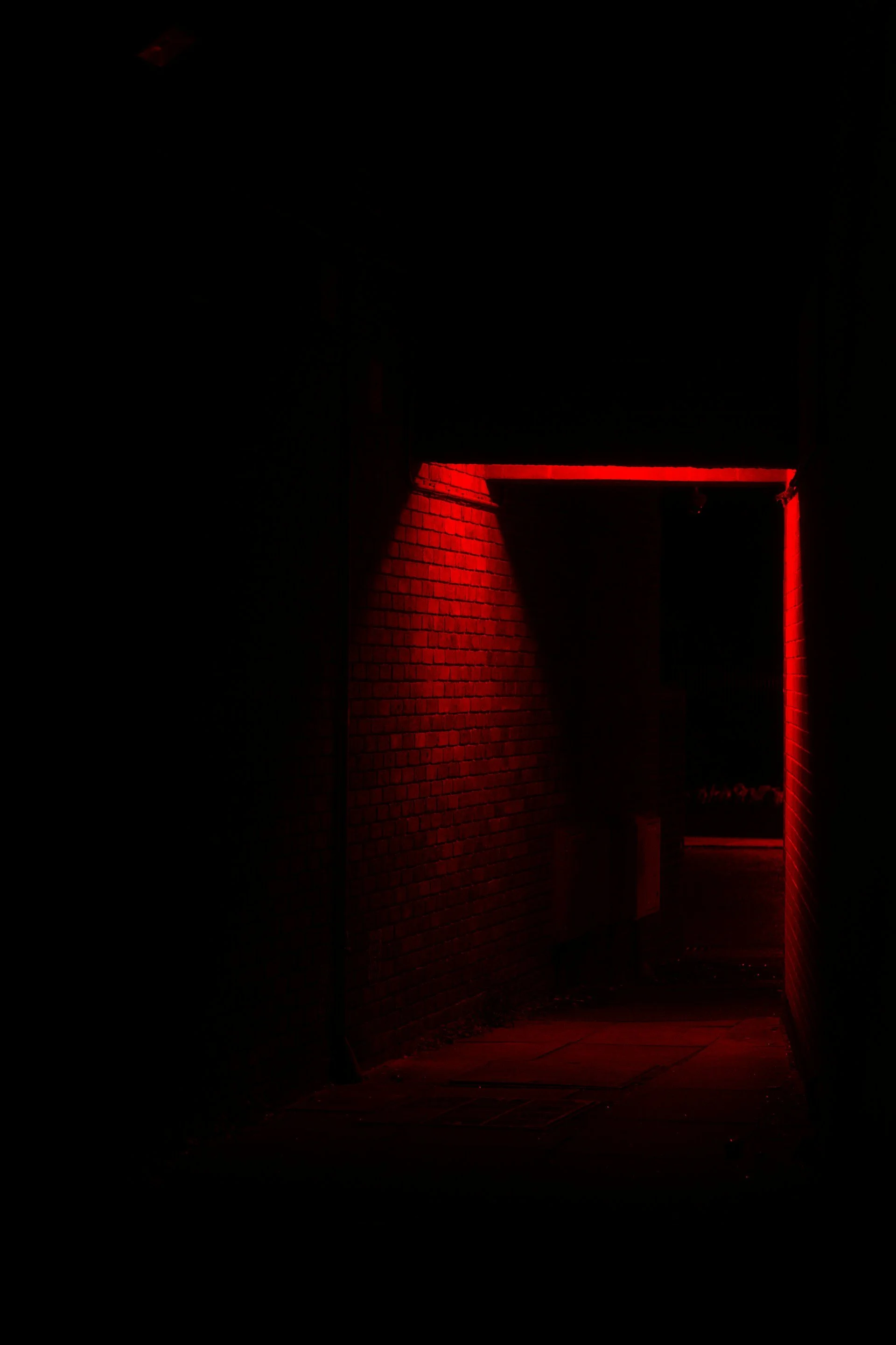 Dark alleyway illuminated by red neon lights along brick walls, leading into darkness.