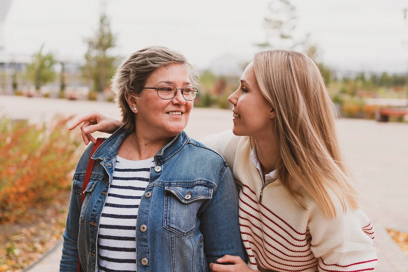 Two women smiling at each other outdoors, one with short gray hair and glasses wearing a denim jacket, and the other with long blonde hair wearing a beige sweater with red stripes.