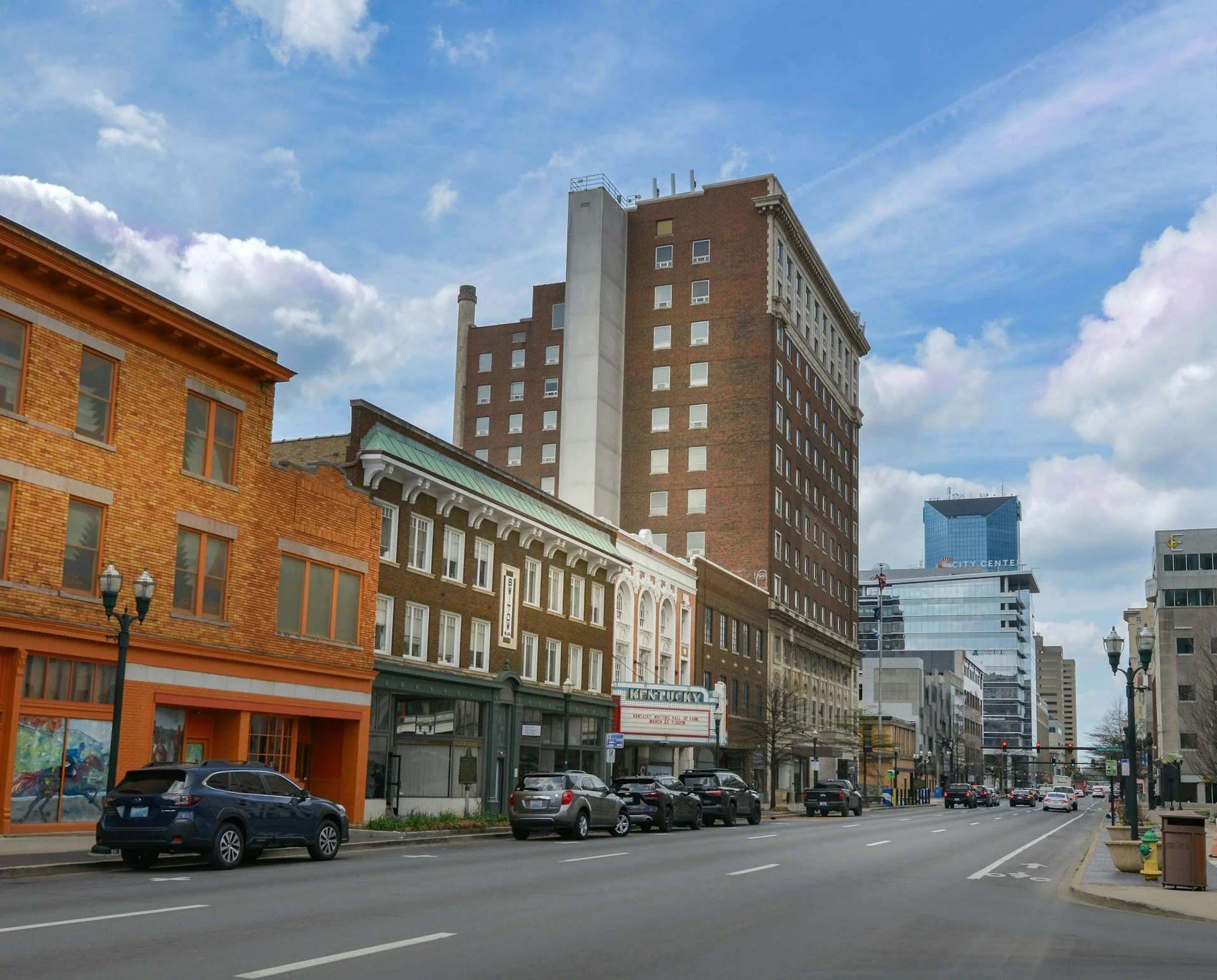 A city street with cars parked along the curb and buildings of various architectural styles. A tall brick office building with many windows is in the background under a partly cloudy sky.
