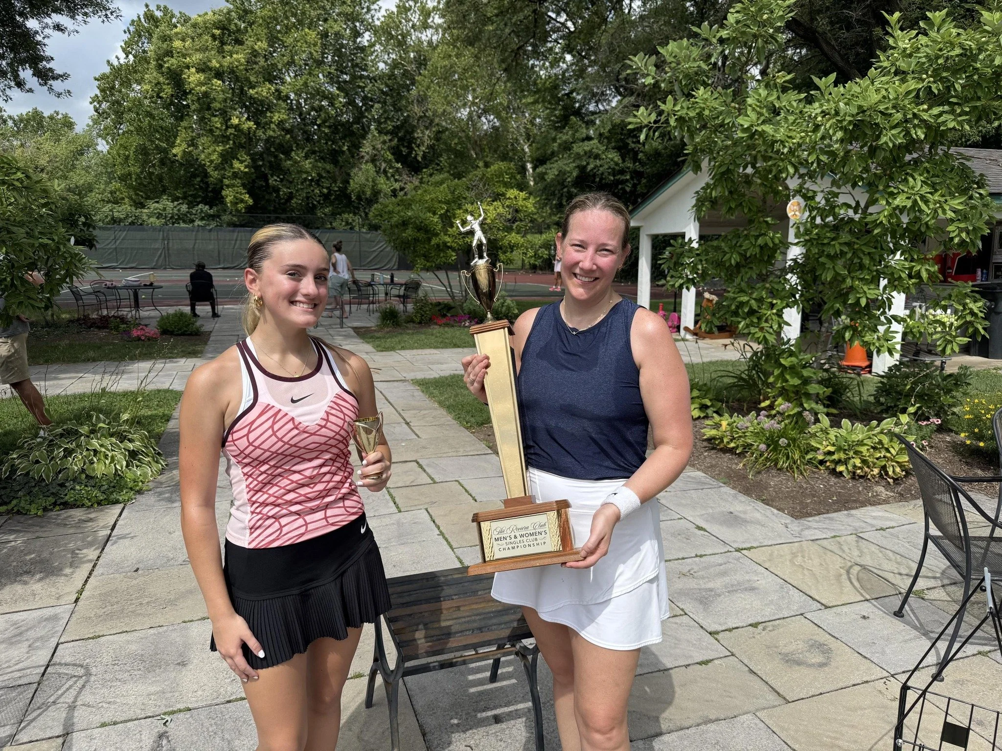 Two women smiling, one holding a trophy, standing outdoors on a patio surrounded by greenery, with tennis courts and people in the background.