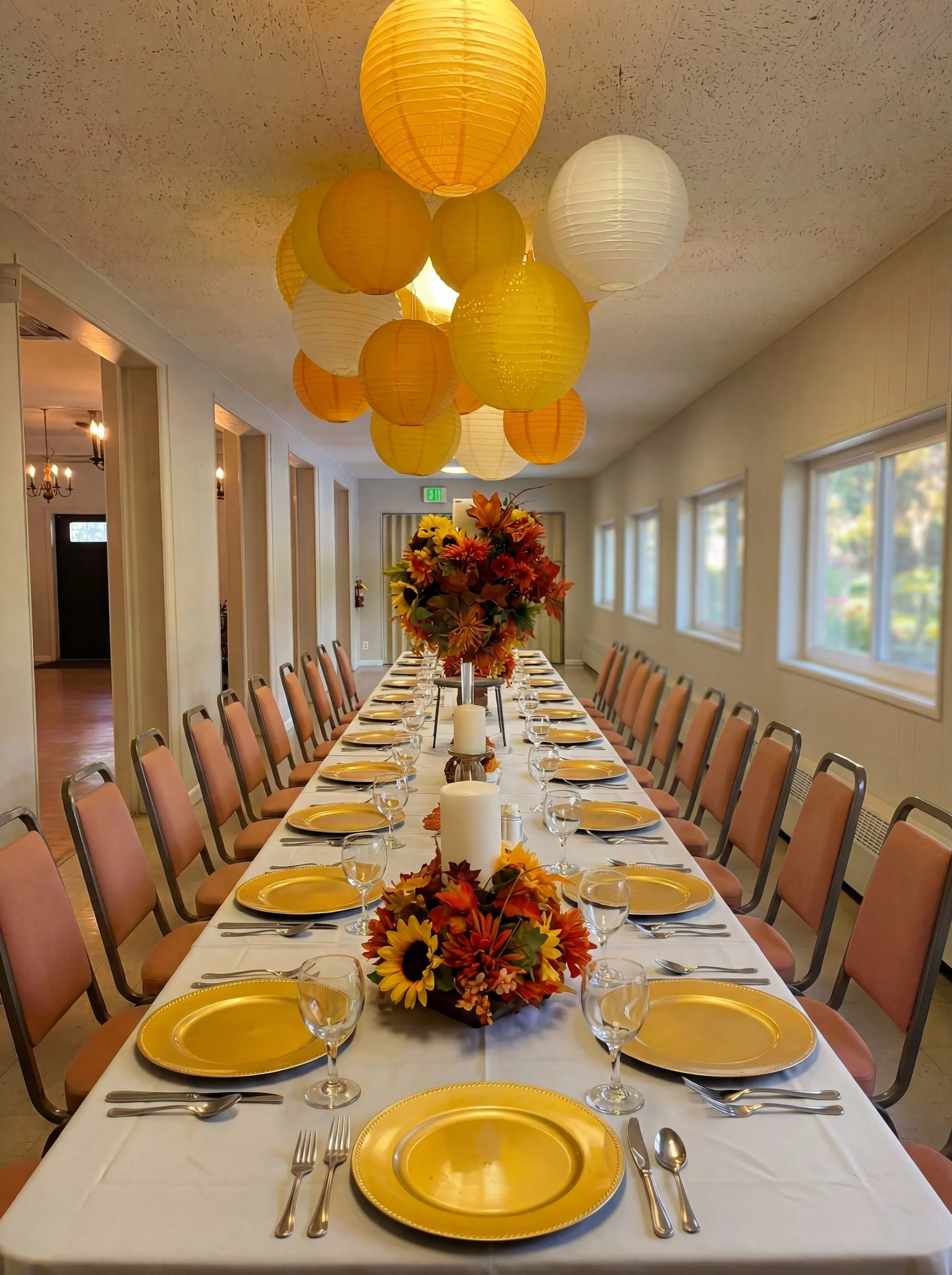 Long dining table with place settings for a formal event, decorated with a fall-themed centerpiece of flowers and leaves, surrounded by beige chairs, with yellow and white paper lanterns hanging from the ceiling.