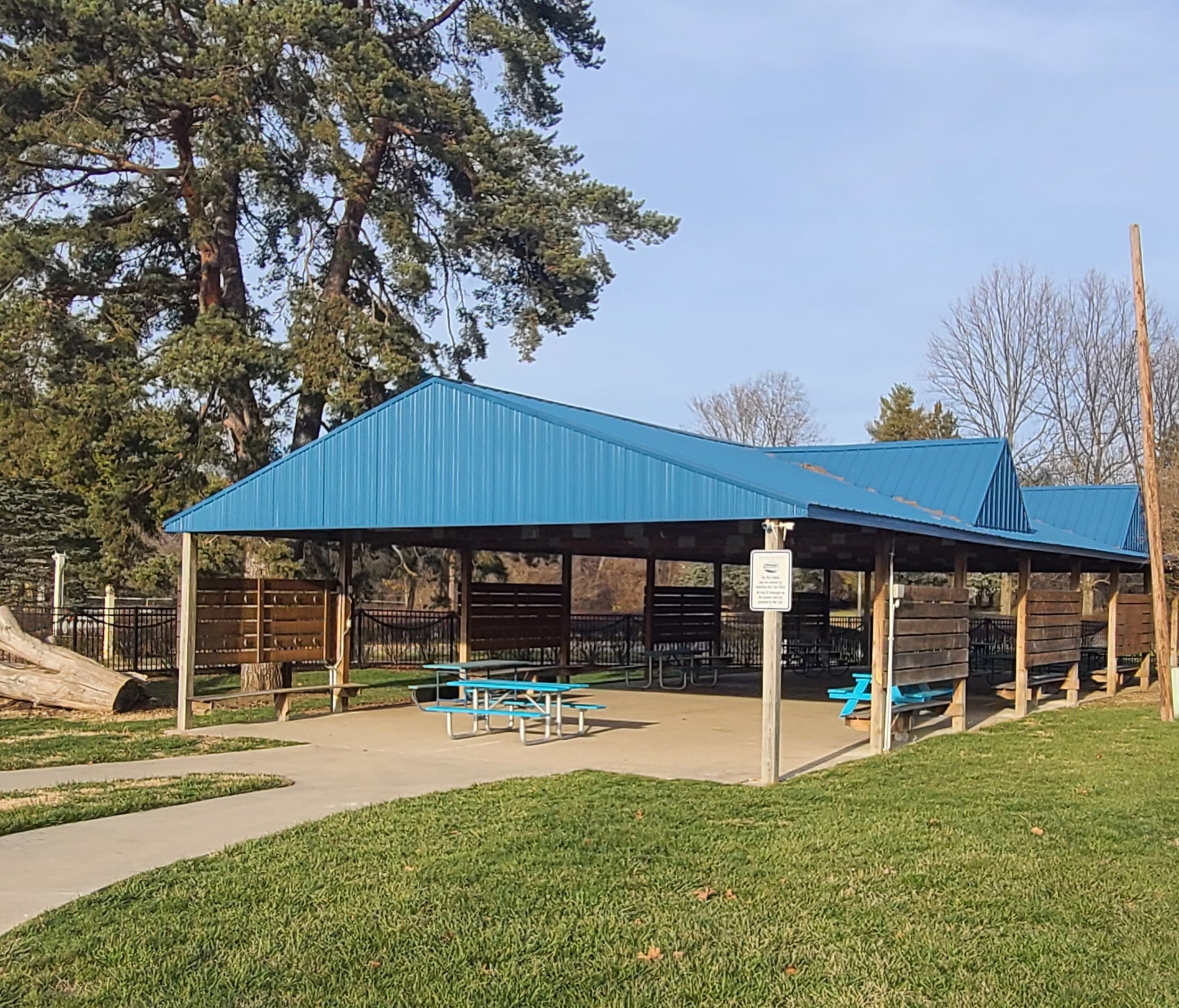 A park area with a blue-roofed pavilion, picnic tables, and a sign in front. Tall trees with some bare branches and a wooden fence surround the area. The sky is clear and blue.