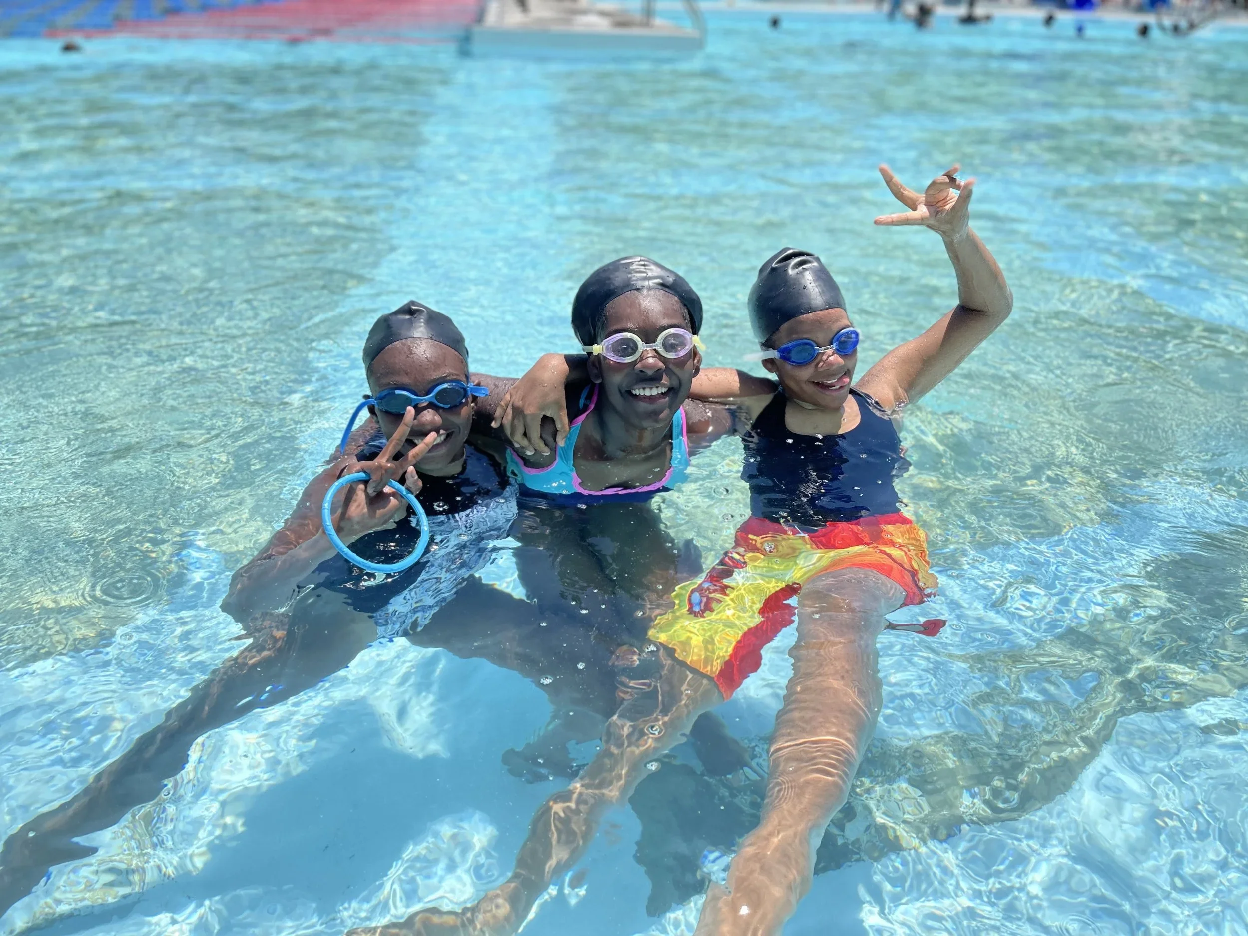 Three children wearing black swim caps and goggles in a swimming pool, smiling and posing for the camera. One child is making a peace sign with their fingers.