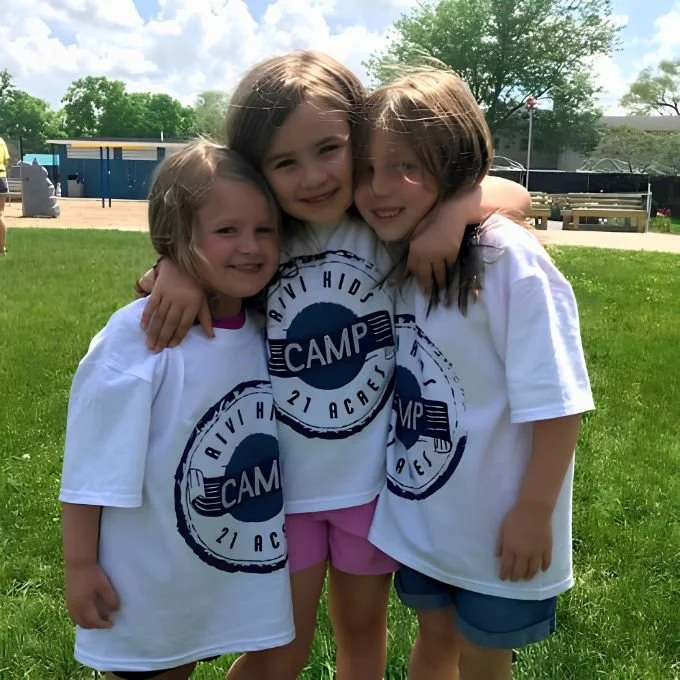 Three children hugging and smiling outdoors on a grassy field during a sunny day, wearing matching white camp T-shirts.