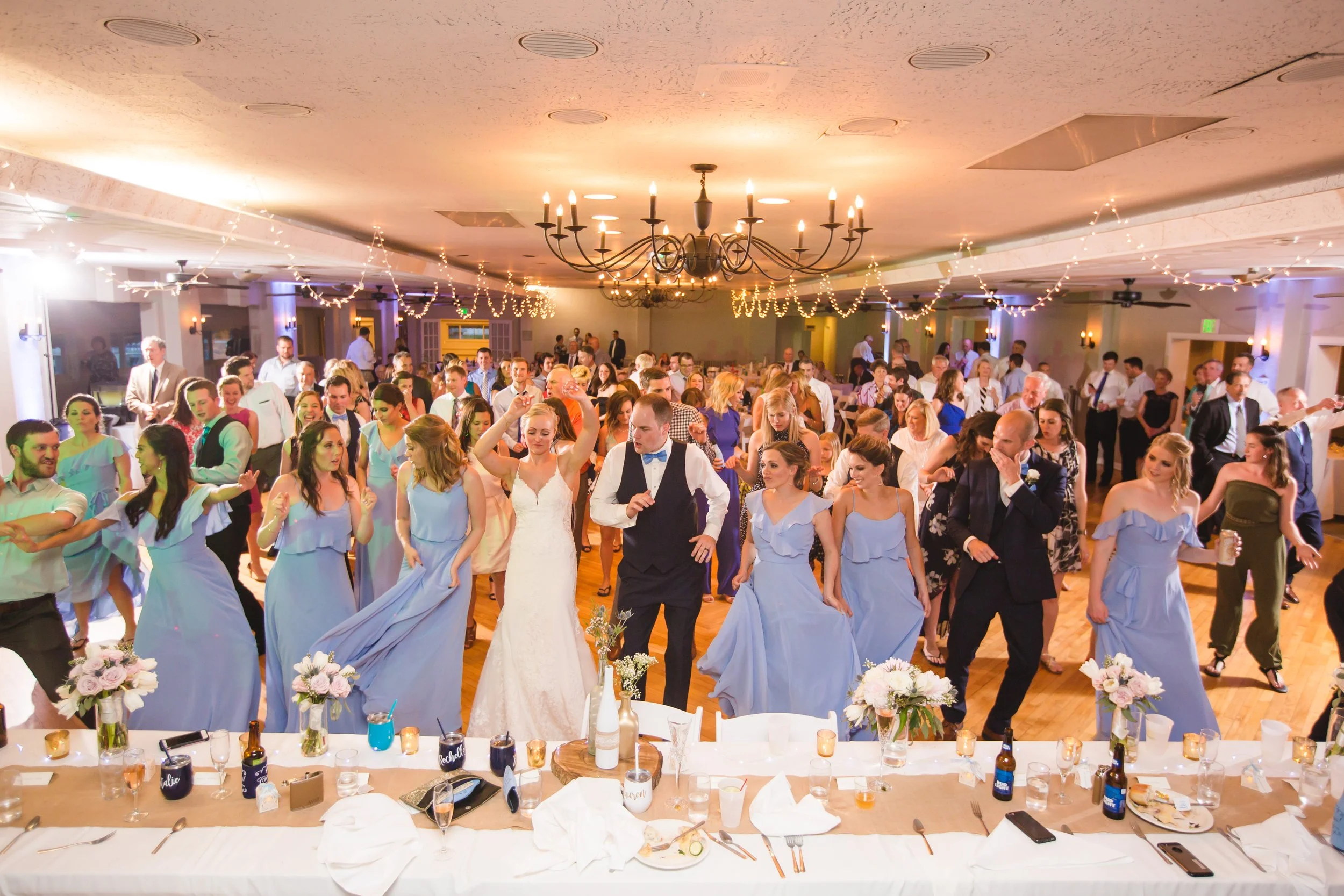 People dancing at a wedding reception with tables, floral arrangements, and string lights in a decorated banquet hall.