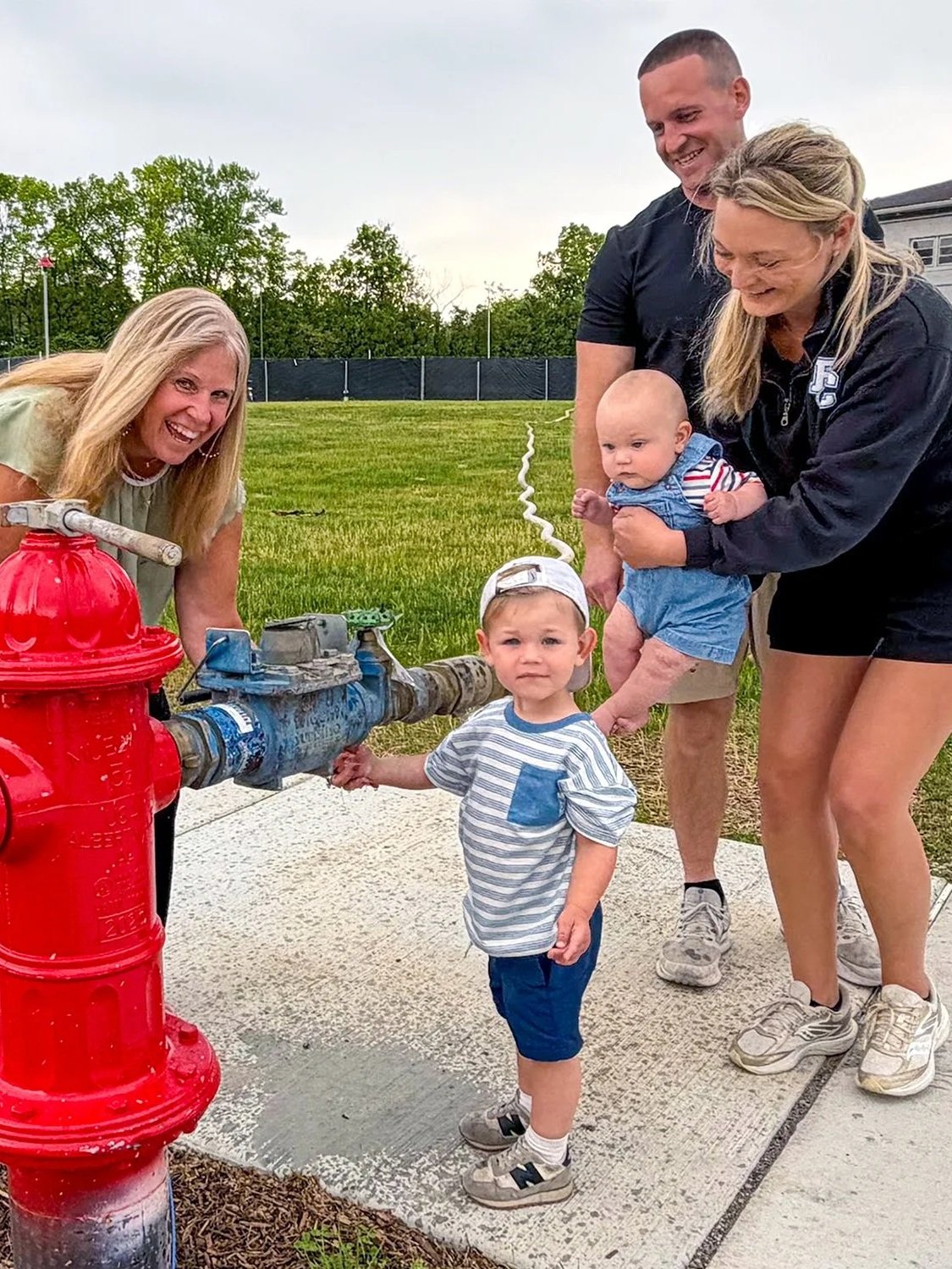 Family of five, including a young boy turning on water at a fire hydrant while a woman holds a baby and a man stands behind them on a grassy field.