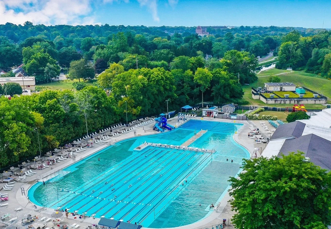 An outdoor swimming pool with multiple lanes and a waterslide, surrounded by lounge chairs and umbrellas, with a lush green forest in the background.