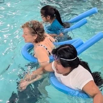 Three women practicing water safety or swim training with pool noodles in a swimming pool.