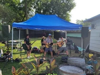 A small outdoor band performance under a blue canopy with five musicians, one playing guitar, others singing and playing instruments, in a backyard with trees and a house in the background.