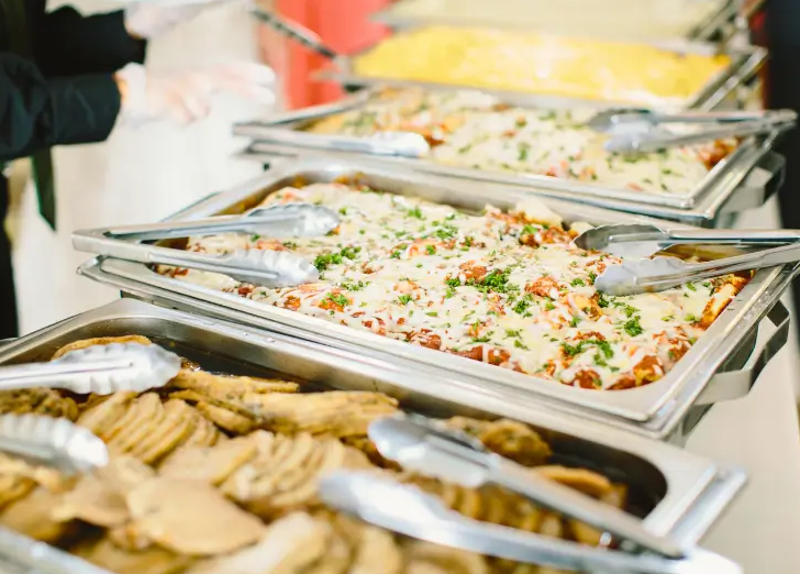 Buffet table with trays of pizza, cookies, and possibly scrambled eggs, with serving tongs.