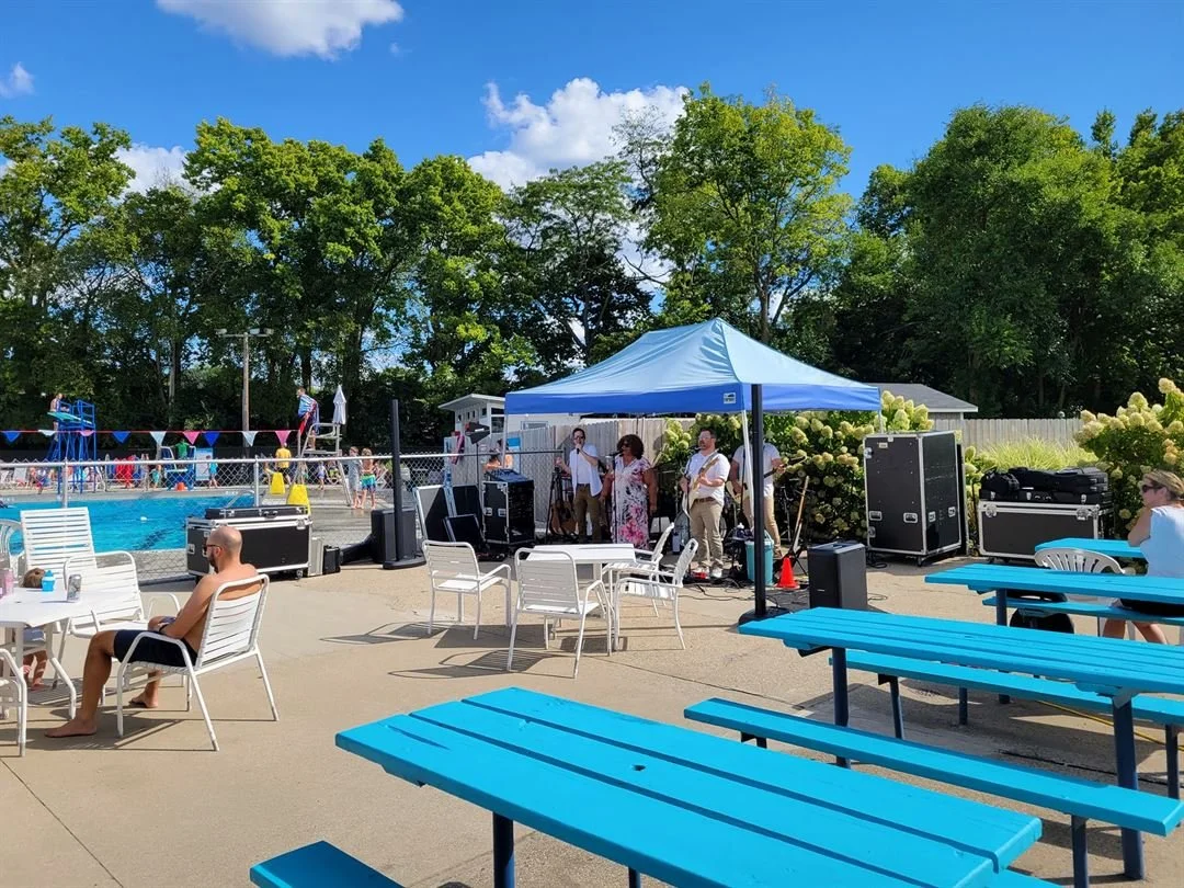 People at a swimming poolside event with a live band performing under a blue canopy, while others relax on chairs and benches on a sunny day. The pool has a lifeguard stand and swimmers in the background, surrounded by green trees.