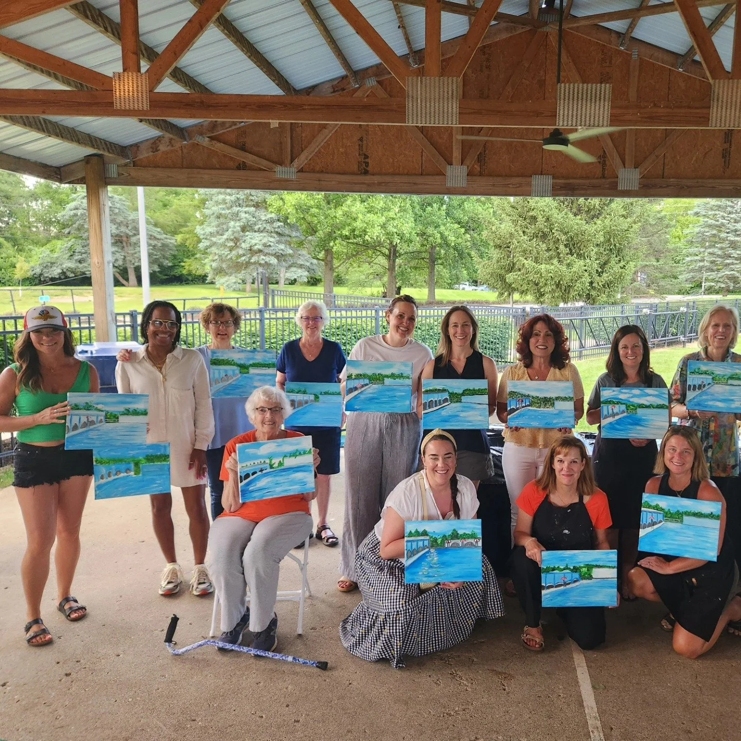 Group of women at an outdoor painting class holding paintings of a bridge over water with trees and sky in the background.