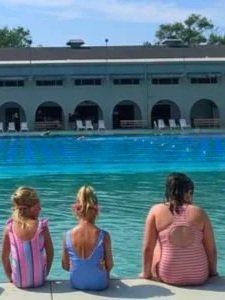 Three young girls sitting by the edge of a swimming pool, facing the water, with a building and blue sky in the background.