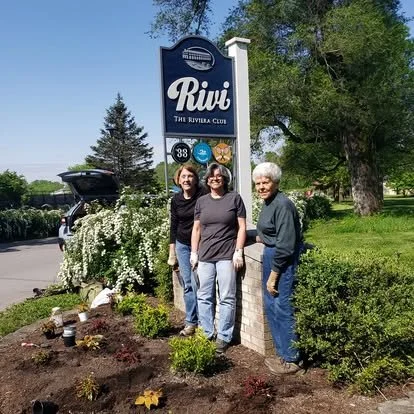 Three women standing near a sign for Rivvi The Riviera Club, in a landscaped garden with trees and bushes, on a sunny day.