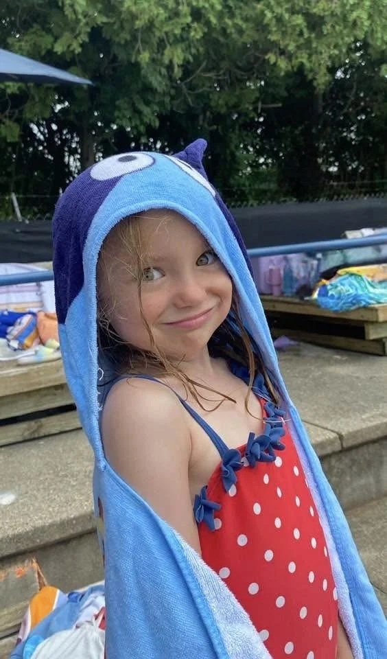 A young girl in a red polka dot swimsuit and a blue towel with a cartoon character hood, smiling outdoors with trees and a picnic table in the background.