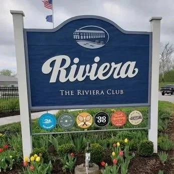 A large blue and white sign for Riviera: The Riviera Club, with several circular logos below it, surrounded by flowers and greenery.