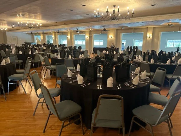 A banquet hall set up for a formal event with round tables covered in black tablecloths, neatly arranged with white napkins, cutlery, and glasses. Chairs surround each table, and the room features chandeliers and large windows letting in natural light.