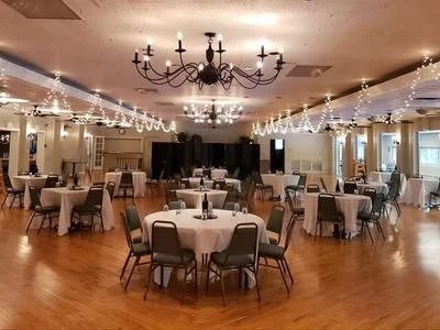 A banquet hall decorated for an event with round tables covered in white tablecloths and surrounded by chairs, featuring string lights and a chandelier on the ceiling.