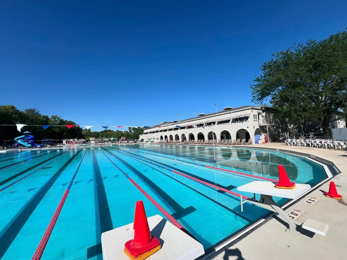 Empty outdoor swimming pool with lane markers, orange traffic cones, and a multi-story white clubhouse in the background under a clear blue sky.