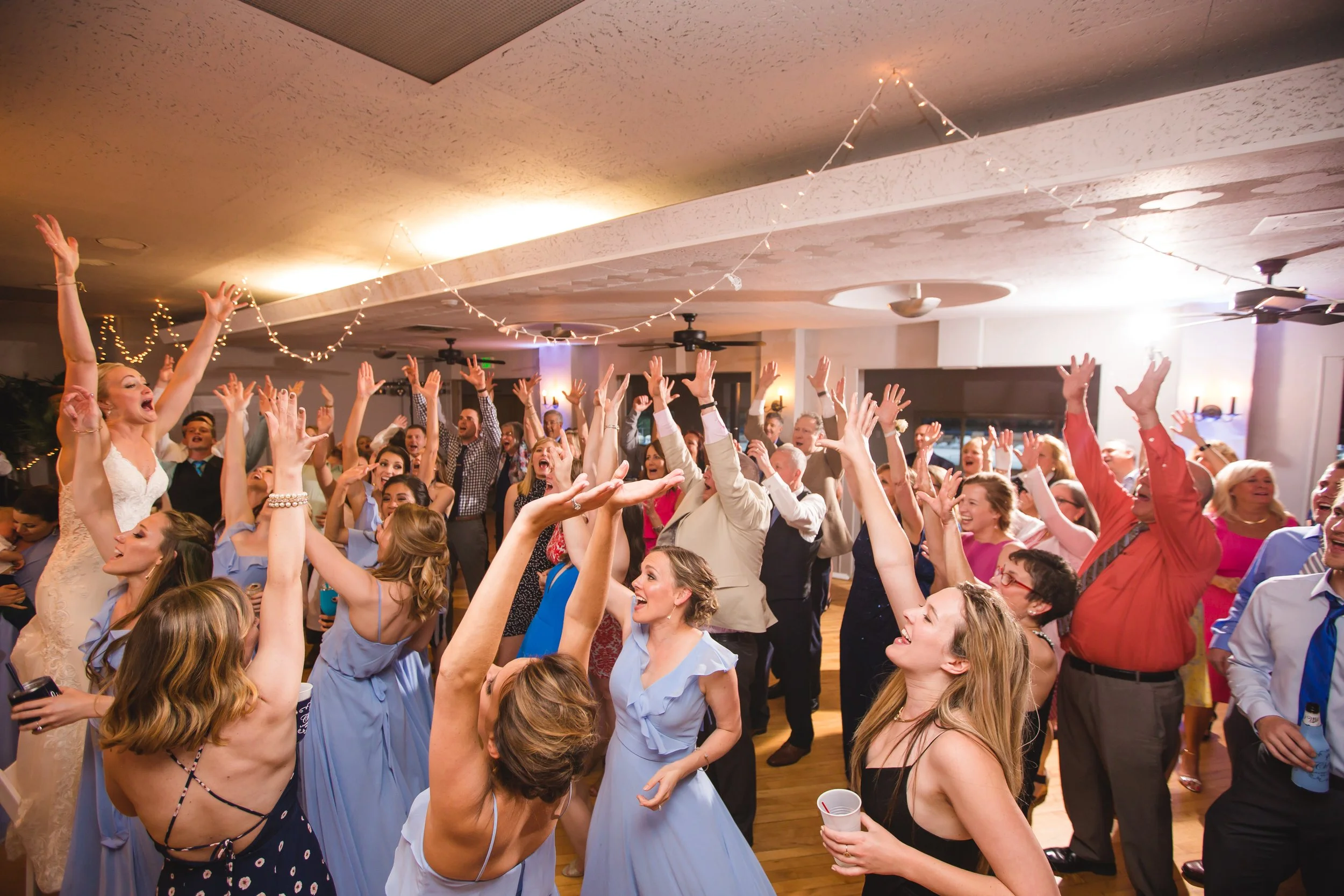 A joyful group of people dancing and celebrating at a party or wedding reception indoors, with string lights hanging from the ceiling.