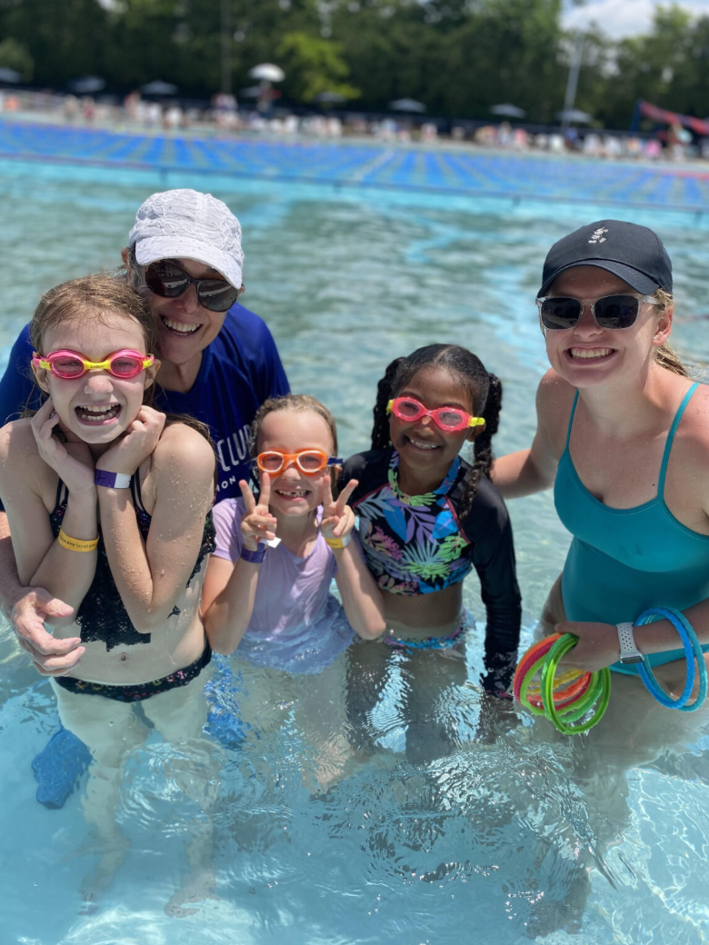 Group of five smiling people, including children and adults, in a swimming pool during a sunny day, wearing swim goggles and swimsuits.