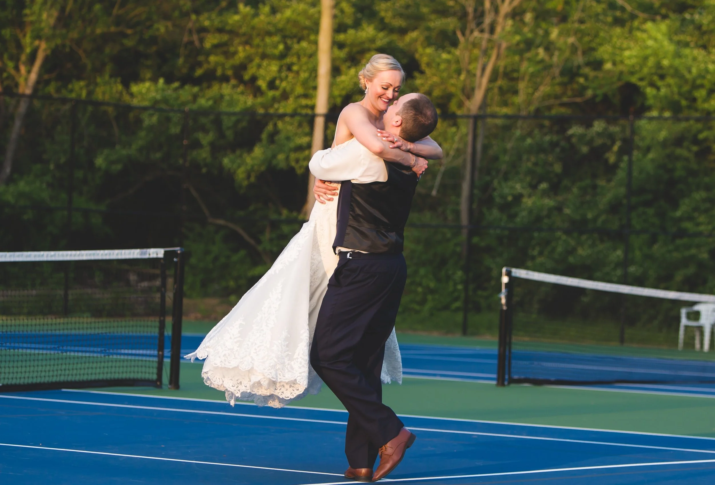 A newlywed couple dancing on a tennis court outdoors, with trees in the background. The bride is wearing a white wedding dress, and the groom is dressed in a dark vest and pants. The bride is smiling, and they are embracing each other.