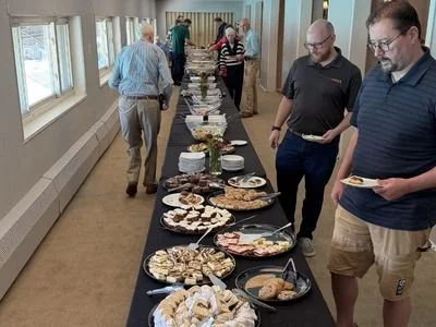 People standing around a long table with various plates of food, such as cookies, desserts, and appetizers, in a bright room with large windows.