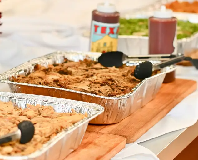 Dinner spread with two aluminum trays of pulled pork, barbecue sauce, and condiments on a wooden board at a picnic.