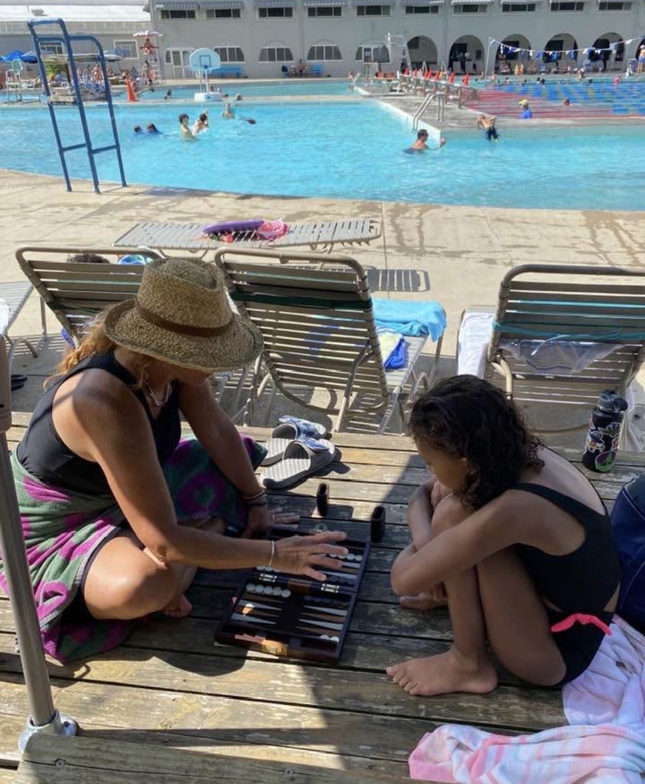 Two women sitting on a wooden poolside deck, one wearing a straw hat and colorful towel, and the other with curly hair, both watching a game on a board. Behind them, people swim and relax at a clear blue outdoor swimming pool, with pool chairs and umbrellas around.