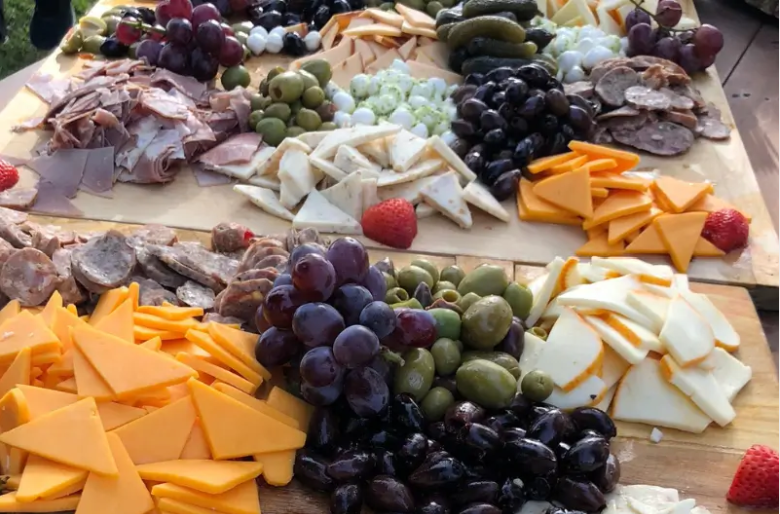 A cheese and charcuterie board featuring sliced Cheddar cheese, sliced mozzarella, green and purple grapes, black olives, green olives, sliced cured meats, and strawberries.