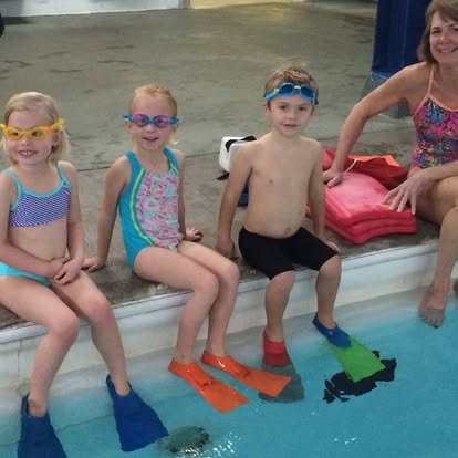Three children and one adult sitting on the edge of a swimming pool, wearing goggles and flippers, with a woman smiling nearby.