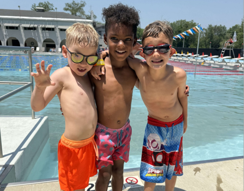 Three young boys in swimsuits standing together at a swimming pool, smiling and hugging each other.