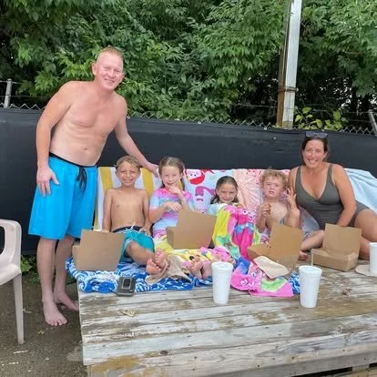 Family enjoying a backyard gathering with kids sitting on a picnic table, opened boxes, and drinks, with two adults standing and smiling.