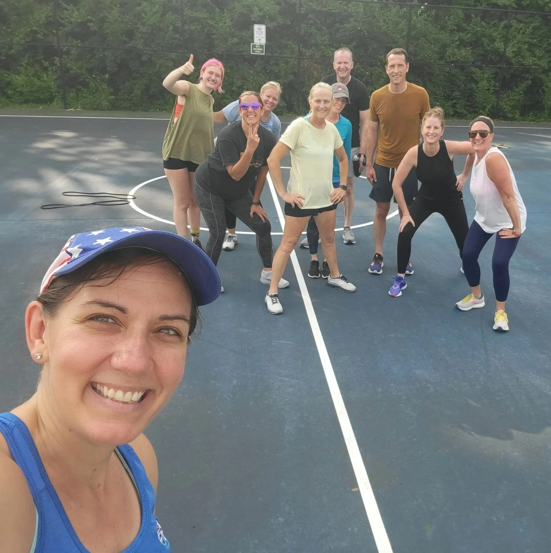 Smiling woman in blue athletic top taking selfie with a group of people on an outdoor basketball court surrounded by trees.