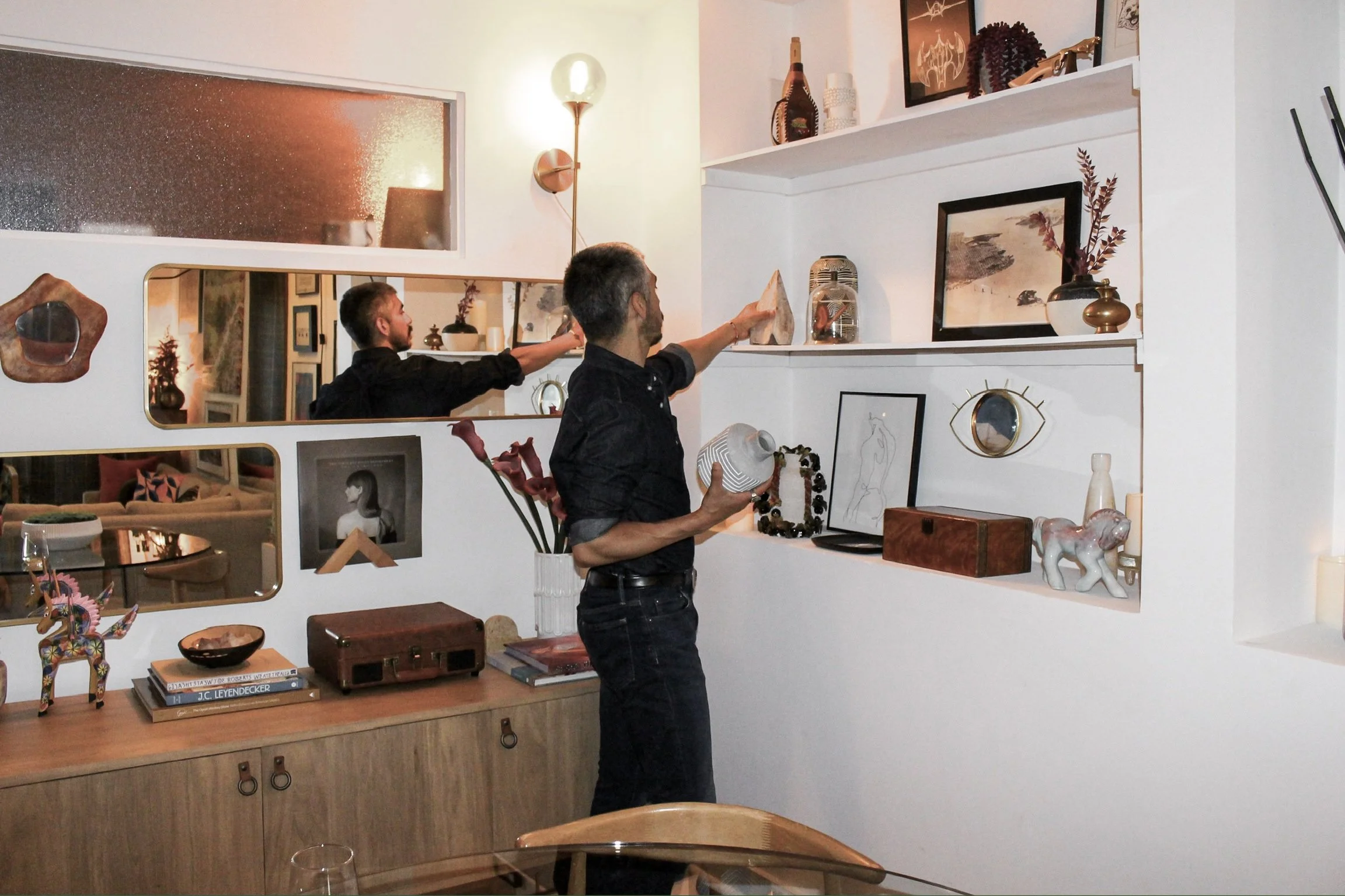Man holding (Adrian Jacob) a patterned vase, standing in a room with artwork and decorative objects on white shelves, mirror, and a wooden console table with books and ornaments.